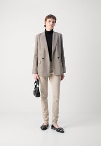 Brun rutigt blazer med dubbla knappar, svart polotröja, beige byxor och svarta flats med rosett. Svart handväska i handen.