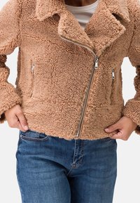 Persona che indossa una giacca in sherpa color beige con zip asimmetrica e jeans blu, mani appoggiate sui fianchi.