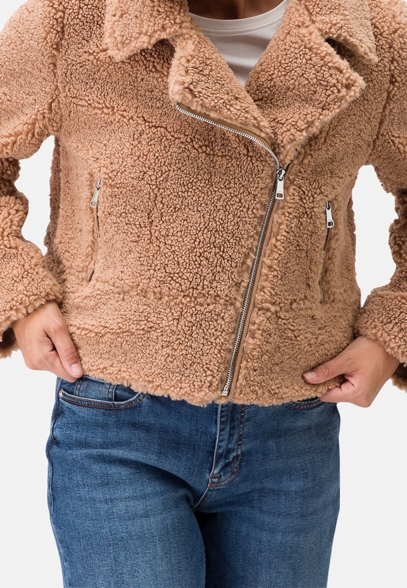 Persona che indossa una giacca in sherpa color beige con zip asimmetrica e jeans blu, mani appoggiate sui fianchi.