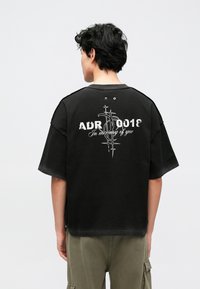 Μαύρο υπερμεγέθες t-shirt με στρογγυλή λαιμόκοψη. Διαθέτει λευκό κείμενο και γραφικό στην πλάτη. Κατασκευασμένο από βαμβάκι με κοντά μανίκια και άνετη εφαρμογή.