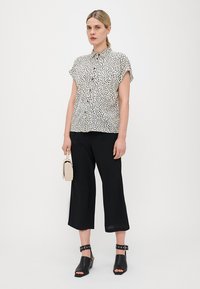 Une chemise à manches courtes avec un motif à pois noir et blanc ; associée à un pantalon noir à jambes larges et des sandales à talons noires.
