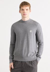 Pulover gri din tricot, cu un guler rotund cu nervuri, textură subtilă și tiv ajustat, având un mic patch cu logo pe piept.