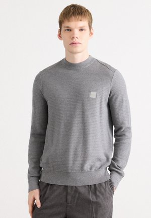 Grauer Strickpullover mit einem gerippten runden Halsausschnitt, subtiler Textur und tailliertem Saum, mit einem kleinen Logo-Patch auf der Brust.