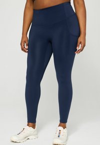 Navy leggings made from a smooth, stretchy fabric, featuring a high waistband and side pockets, paired with white athletic shoes.