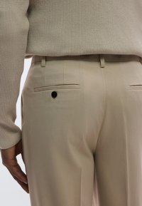 Beige skreddersydde bukser med glatt tekstur, med en enkelt baklomme med svart knapp. Midjebåndet er flatt og rent.