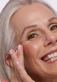 Nærbilde av en smilende person med lys grått hår som påfører krem på kinnet. Huden viser tekstur og subtile rynker; neglene er lyse rosa.