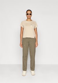 Μπεζ κοντομάνικο μπλουζάκι με τσέπη στο στήθος, σε συνδυασμό με ελιόχρωμους παντελόνες. Λευκά παπούτσια ολοκληρώνουν την εμφάνιση. Το φόντο είναι καθαρό.