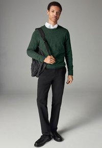 Pull vert foncé, chemise blanche en dessous, pantalon noir et chaussures noires. L'homme porte un sac noir texturé, il est adossé à un fond gris.