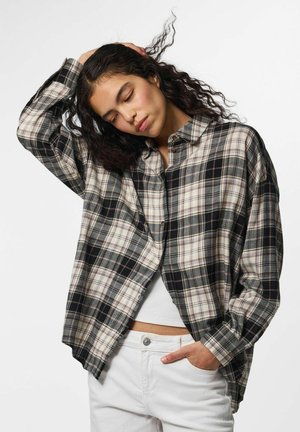 Jeune femme aux cheveux bouclés portant une chemise à carreaux noire et blanche, un crop top blanc et un pantalon blanc, se tenant debout, les yeux fermés et la main sur la tête.