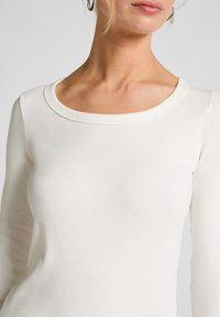 Vrouw in een effen witte lange mouwen geribbelde T-shirt met een ronde hals en zilveren oorringen, weergegeven van nek tot taille.