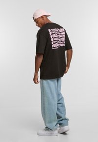 T-shirt nera oversize con grafiche rosa sulla schiena, abbinata a jeans baggy azzurri e sneakers bianche. Cappellino rosa indossato al contrario.