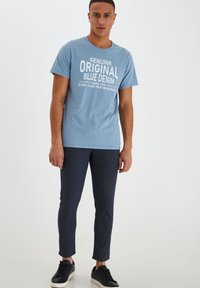 Svijetloplava pamučna majica s natpisom "Genuine Original Blue Denim", u kombinaciji s tamnim teksturiranim uskim hlačama i crnim tenisicama.