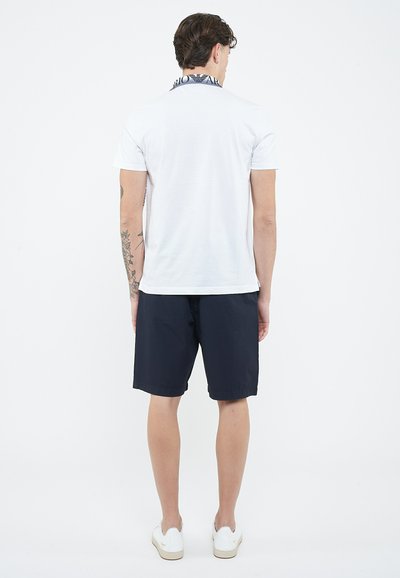Man with tattoos wearing a white polo shirt with patterned collar, black shorts, and white sneakers, standing facing away on plain background.