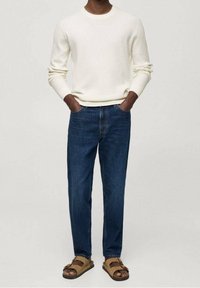 Pull en maille côtelée blanche à manches longues, jeans en denim bleu et sandales marron. La tenue affiche une coupe décontractée et un style casual.