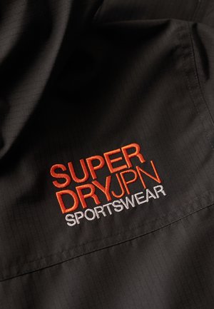 Tessuto nero con una trama a rete, caratterizzato da un logo ricamato in arancione e bianco che recita "SUPERDRYPN SPORTSWEAR" ben in vista.