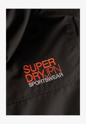 Tessuto nero con una trama a rete, caratterizzato da un logo ricamato in arancione e bianco che recita "SUPERDRYPN SPORTSWEAR" ben in vista.