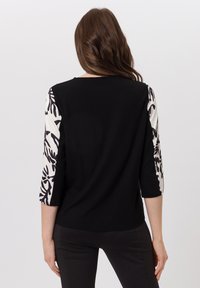 Zwarte top met driekwart mouwen in een witte stof met zwarte abstracte bloemenpatronen, ronde halslijn en een zachte textuur.