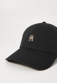 Svart baseball cap laget av teksturert stoff. Har en rund krone, buet skjerm og en liten gulllogo foran.
