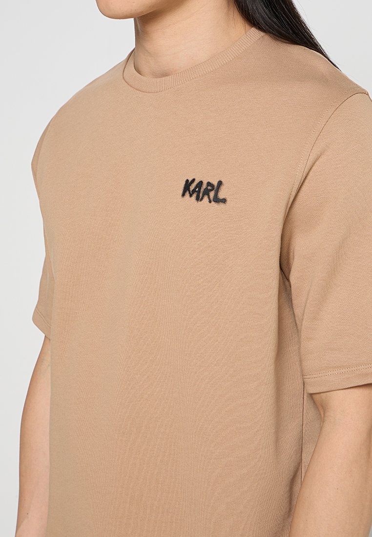 Beżowa koszulka z krótkim rękawem wykonana z bawełny, z czarnym haftowanym logo "KARL" na lewej stronie klatki piersiowej. Gładka tekstura.