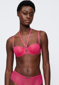 Růžová krajková podprsenka s geometrickým vzorem, vlnitými okraji a nastavitelnými ramínky. Spárována s odpovídajícími růžovými krajkovými kalhotkami se podobným designem.