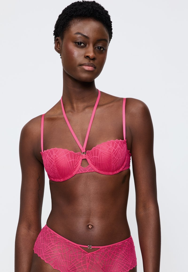Růžová krajková podprsenka s geometrickým vzorem, vlnitými okraji a nastavitelnými ramínky. Spárována s odpovídajícími růžovými krajkovými kalhotkami se podobným designem.