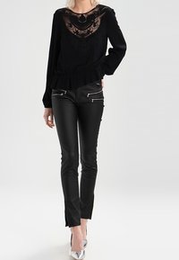 Sort bluse med blonde detaljer og volangkant, paret med sorte tætsiddende bukser med lynlåsdetaljer og sølvhælede sko.