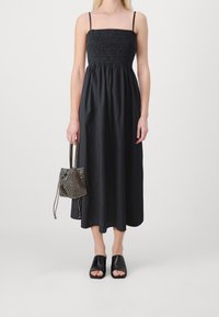 Robe d'été noire à smocks avec des bretelles fines, jupe jusqu'aux genoux et coupe décontractée. Accompagnée d'un petit sac en maille métallique porté en bandoulière et de sandales noires.