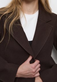 Cappotto in misto lana marrone con rever a punta, spalle strutturate e vestibilità sartoriale. Il modello tiene chiuso il cappotto. Strato interno bianco visibile.