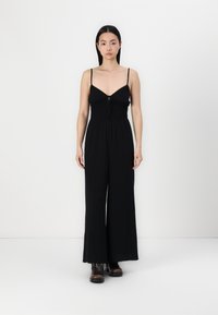 Macacão preto com tiras finas ajustáveis, corpete justo, cintura franzida e calças de perna larga; feito de um tecido leve e suave.