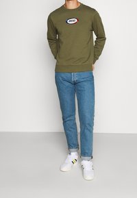 Felpa verde oliva con collo rotondo e logo "DIESEL" ricamato. Abbinata a jeans blu e sneakers bianche con righe colorate.