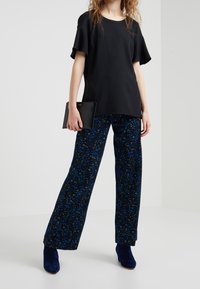 Czarny krótki top z rękawami, zestawiony z granatowymi spodniami w kwiaty o szerokich nogawkach, z niebieskimi i zielonymi wzorami. Modelka trzyma czarną torebkę z teksturowanej tkaniny.
