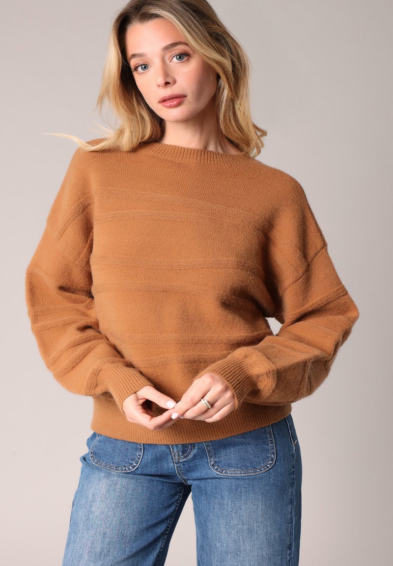 Jeune femme aux cheveux blonds, portant un pull marron ample et un jean bleu, les mains jointes à la taille devant un fond uni.