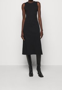 Robe midi noire sans manches cintrée, assortie à des bottes en cuir noir jusqu'aux genoux, portée par une personne debout devant un fond blanc uni.