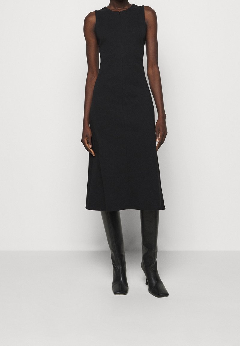 Robe midi noire sans manches cintrée, assortie à des bottes en cuir noir jusqu'aux genoux, portée par une personne debout devant un fond blanc uni.