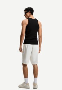 Top fără mâneci, de culoare neagră, pantaloni scurți albi și adidași de culoare deschisă; model cu păr creț și ochelari de soare văzut din spate.