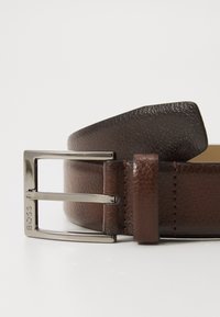 Ceinture en cuir marron avec une finition texturée, dotée d'une boucle rectangulaire en argent et d'un gravage de marque. Détails cousus le long du cuir.