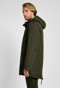 Oliwkowy parka z kapturem, zapinany na zamek z przodu, oraz dwoma bocznymi kieszeniami. Posiada gładką teksturę i regulowany sznurek w dolnej części.