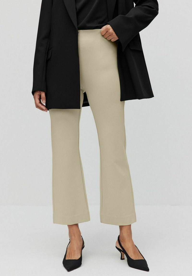 Beige utsvängda byxor i stretchy material med hög midja och sömdetaljer, samt en svart överstor blazer och svarta klackar.