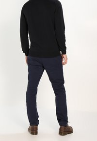 Dunkel gestrickter Pullover mit Rundhalsausschnitt, über einer schmal geschnittenen blauen Hose getragen. Ausgestattet mit einer glatten Textur und Bündchen, kombiniert mit braunen Schuhen.