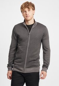 Sudadera gris con cremallera, con un patrón de punto texturado, puños y cintura acanalados, llevada sobre una camiseta negra. Tiene un diseño de cuello alto.