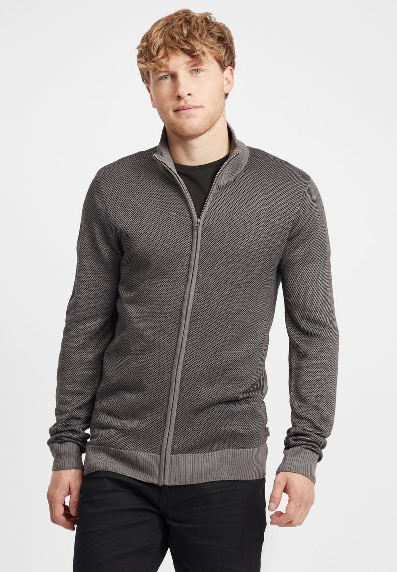Sudadera gris con cremallera, con un patrón de punto texturado, puños y cintura acanalados, llevada sobre una camiseta negra. Tiene un diseño de cuello alto.