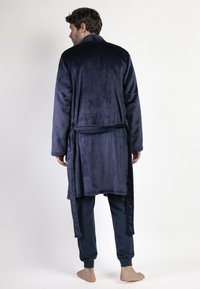 Hombre de pie descalzo, vistiendo un albornoz de felpa azul oscuro con cinturón adjunto y pantalones oscuros a juego, de espaldas contra un fondo blanco.