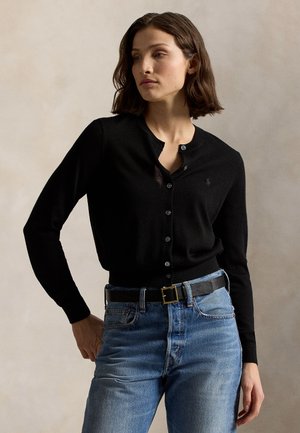 Kvinde iført en knappet sort cardigan, stukket ind i højtaljede blå jeans med sort bælte, stående mod en ensfarvet baggrund.