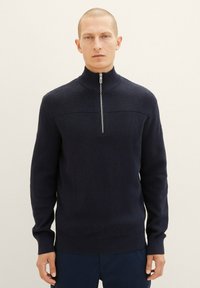 Dunkelblauer gestrickter Pullover mit hohem Kragen, der einen halben Reißverschluss und ein strukturiertes Rippenmuster im oberen Bereich aufweist. Lange Ärmel.