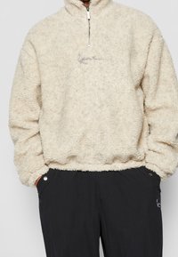 Fleece-Pullover in Creme mit hohem Kragen und halbem Reißverschluss; verfügt über ein dezentes Textlogo auf der Vorderseite und einen elastischen Saum.