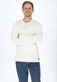 Vit långärmad henleytröja med tre bruna knappar vid halsringningen, tillsammans med blå jeans. Tyget ser texturerat och mjukt ut.