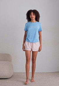 Světle modré bavlněné tričko s logem "Calvin Klein", doplněné o měkké růžové šortky. Model stojí bosý na neutrálním pozadí.