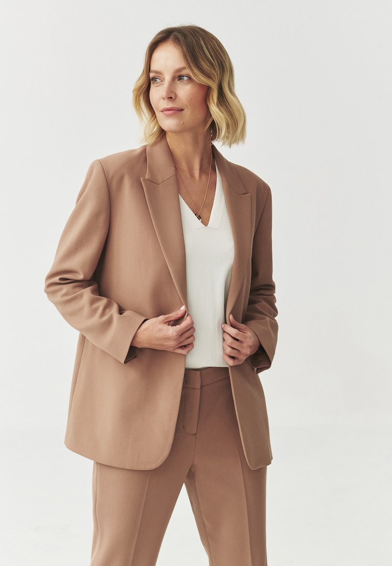 Blazer beige con una vestibilità rilassata, top bianco a scollatura a V sottostante e pantaloni beige coordinati. Texture liscia e design minimalista.