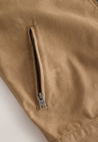 Tissu kaki en gros plan avec une fermeture éclair verticale partiellement ouverte dotée d'un curseur métallique sur une manche de vêtement ou une jambe de pantalon.
