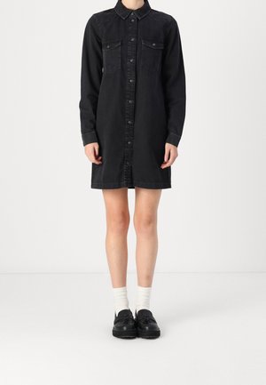 Person iført en sort skjortekjole med lange ærmer, hvide crew-sokker og sorte loafers med kvaster, står mod en hvid baggrund.